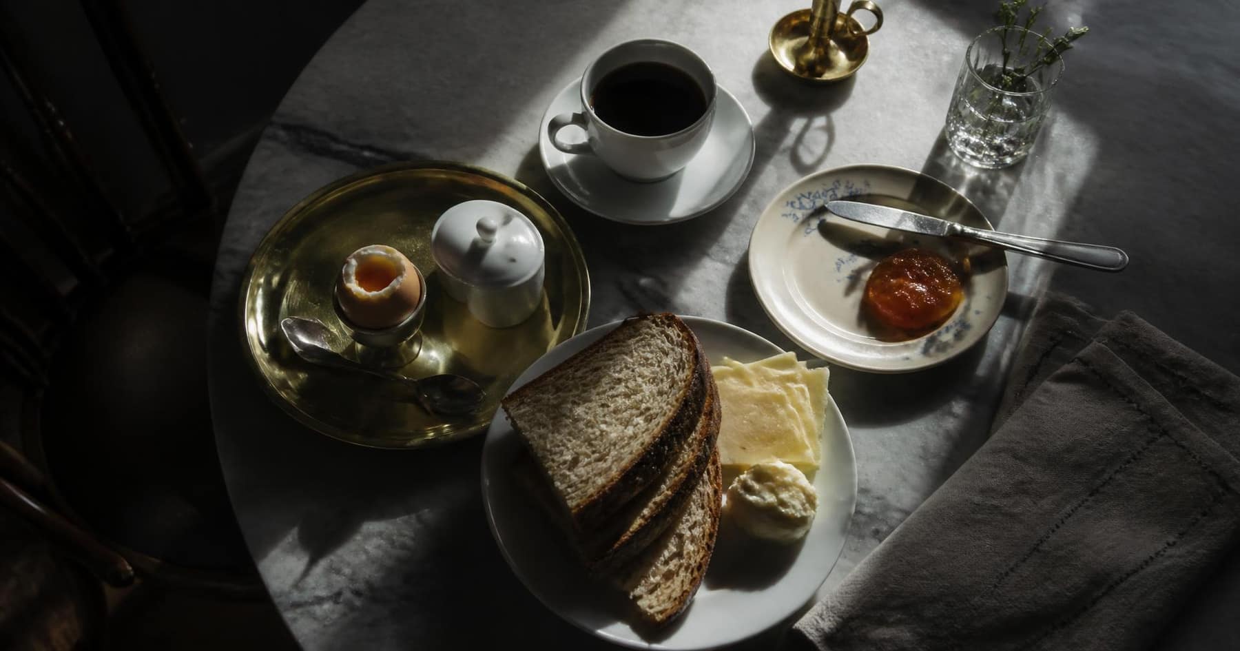 Frukostbricka med kokt ägg, surdegsbröd, ost, marmelad och kaffe på ett marmorbord